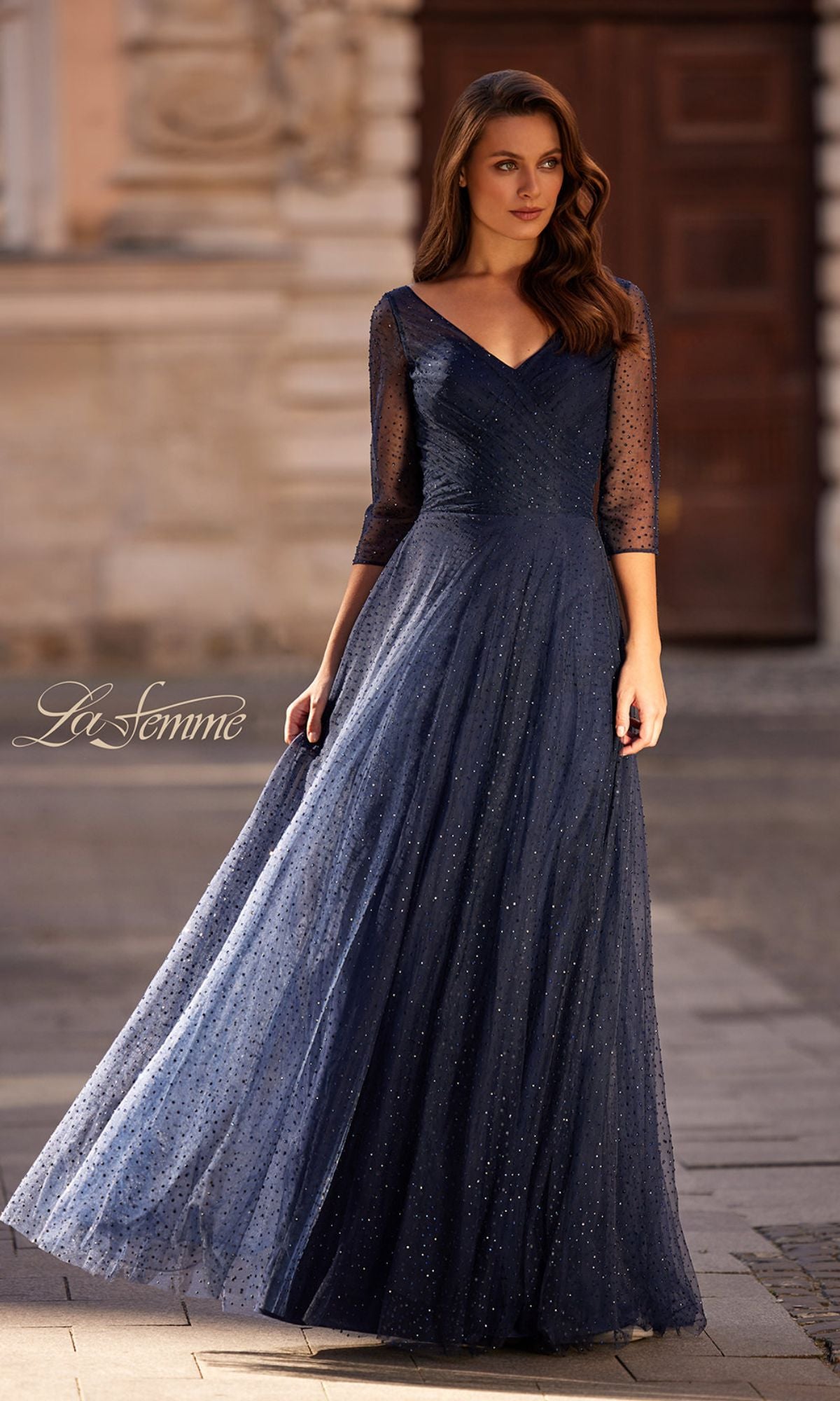 A woman stands outdoors in the La Femme 32460 Sparkly Sleeved Tulle Formal Dress, featuring sheer three-quarter sleeves. A blurred building and door in the background accentuate her elegant, shimmering look.